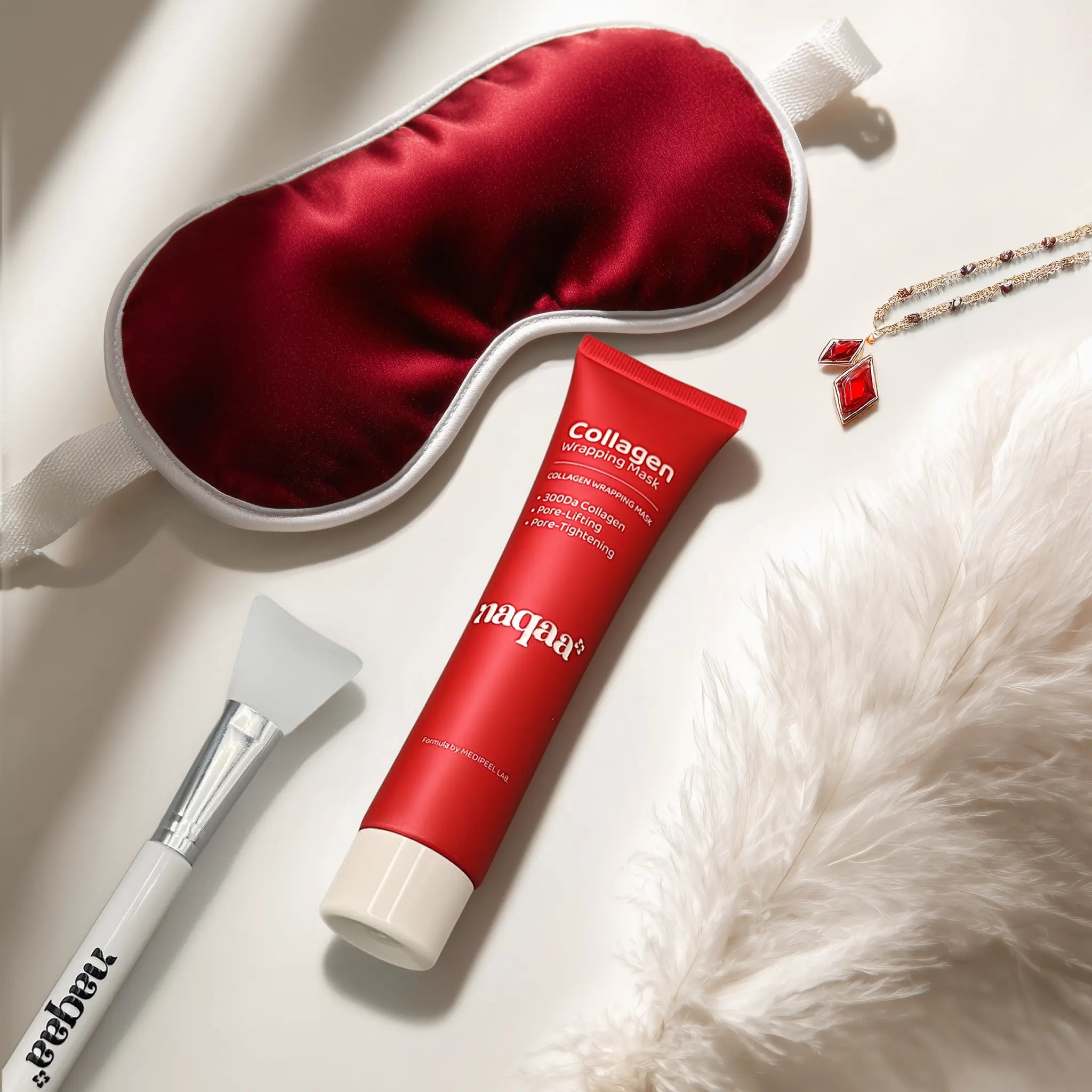 Luxurious flat lay of the Naqaa Collagen Wrapping Mask, applicator brush, and red silk sleep mask for a relaxing self-care routine.